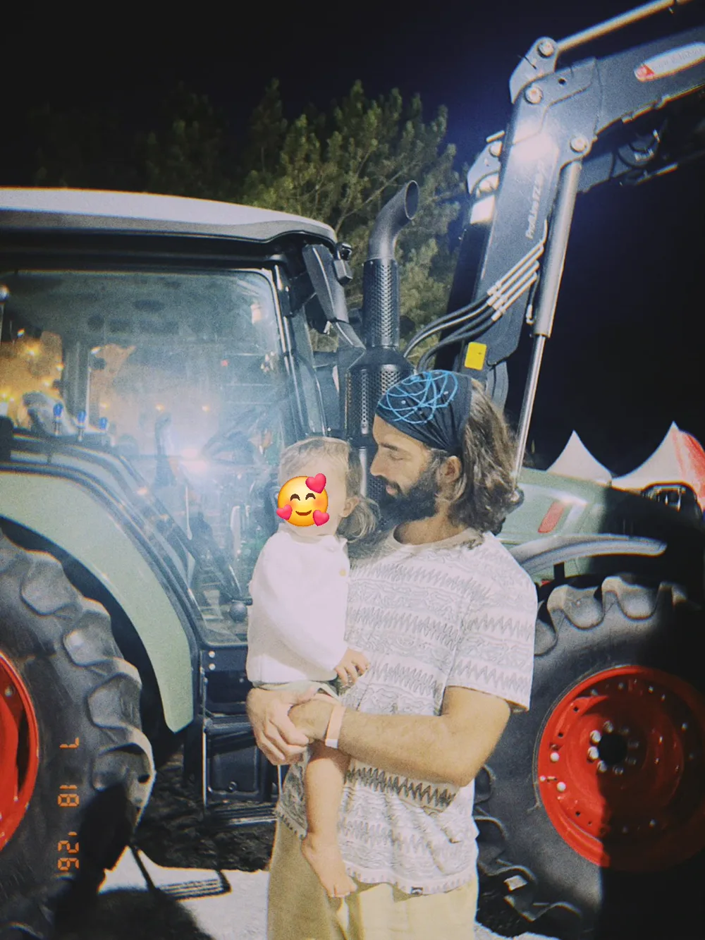 Tiago at night in front of a big green tractor with red wheels, holding his son, whose face is covered with an emoji