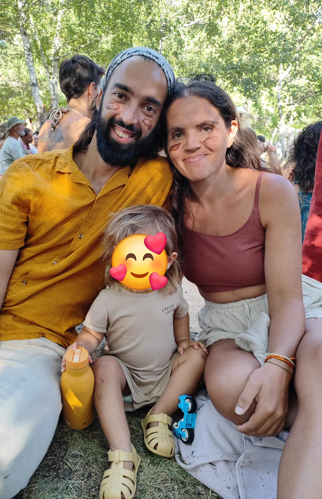 Tiago and Irina sitting on the grass at a gathering with their son between them, his face covered with an emoji