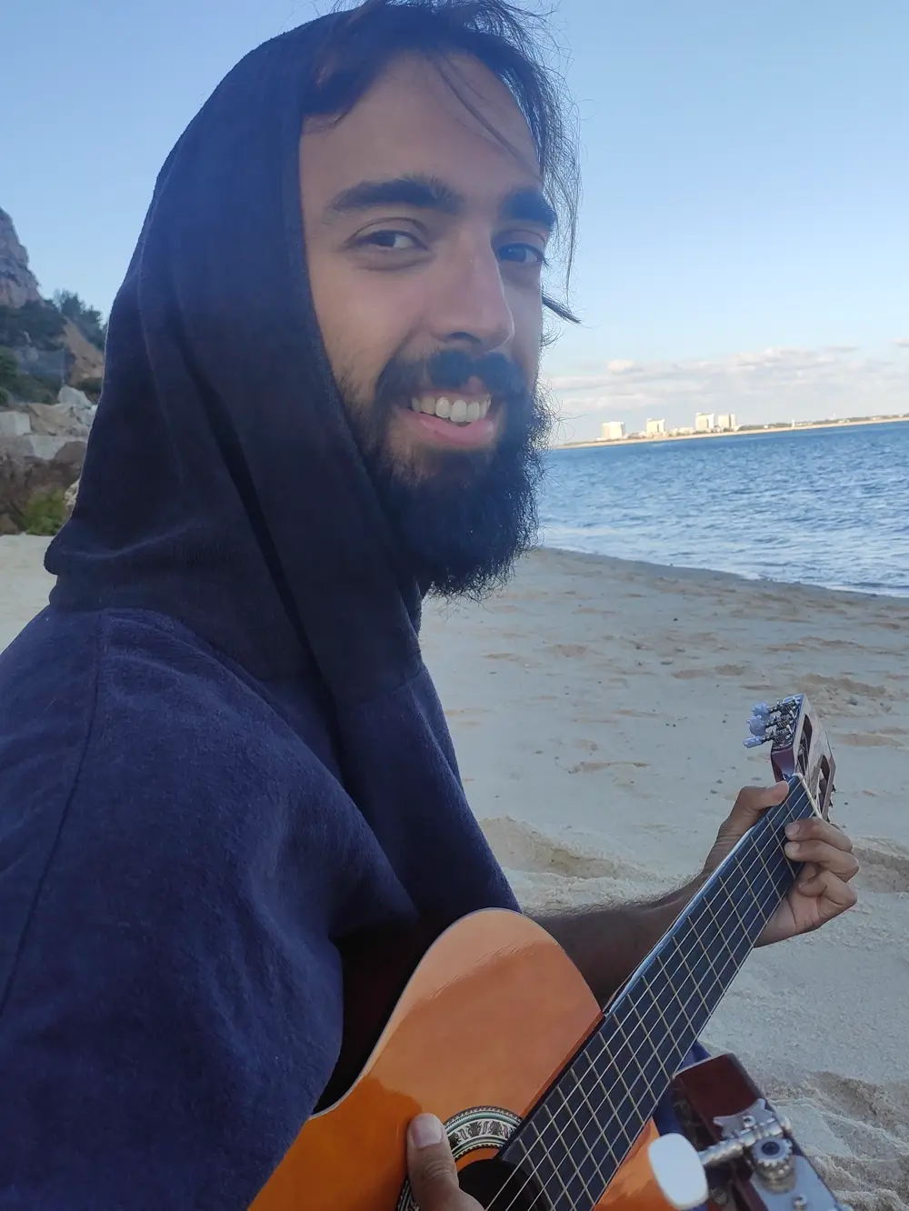 Tiago in a hood on a beach, smiling at the camera with a guitar
