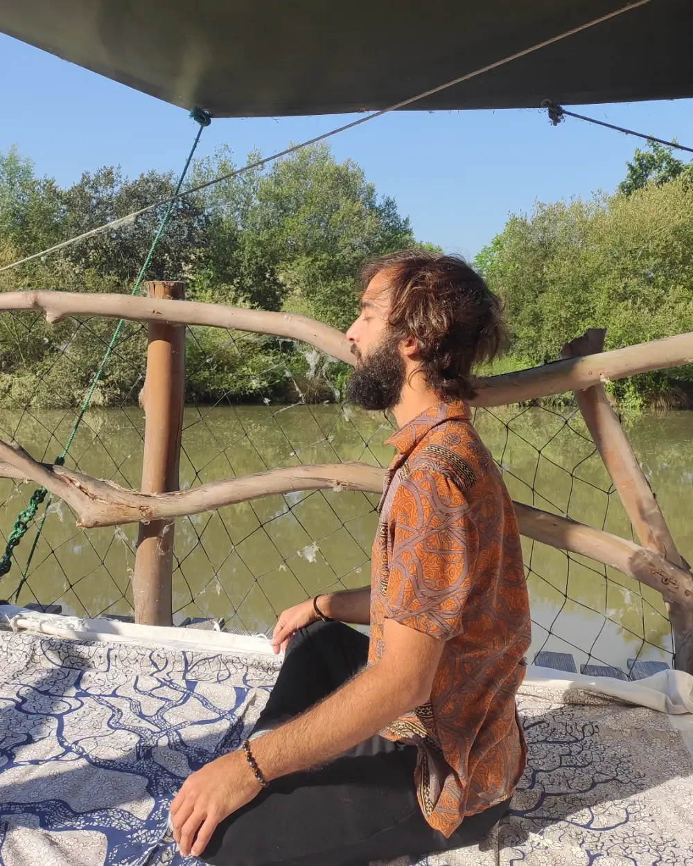 Tiago meditating cross-legged on a wooden deck by a river, in an orange shirt, with trees behind him