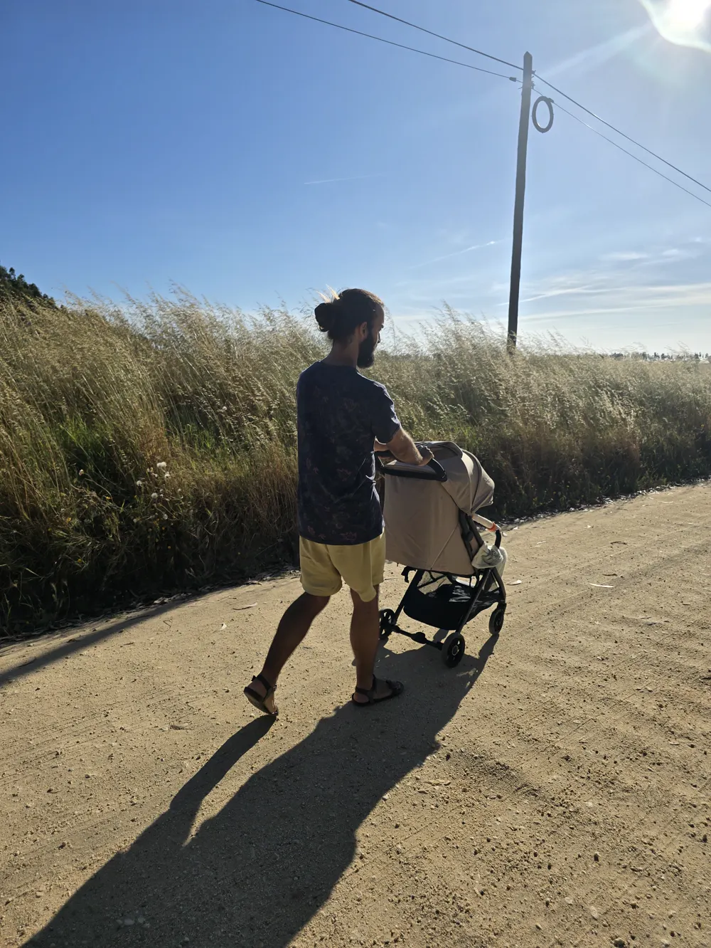 Tiago from behind on a dirt road in the Alentejo, pushing a pram past tall dry grass under a blue sky