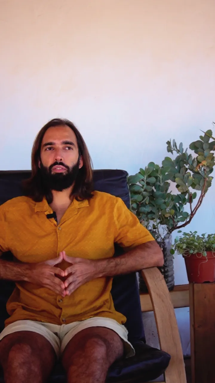 Tiago in a yellow shirt, sitting in a wooden chair on a terrace with plants, talking to the camera